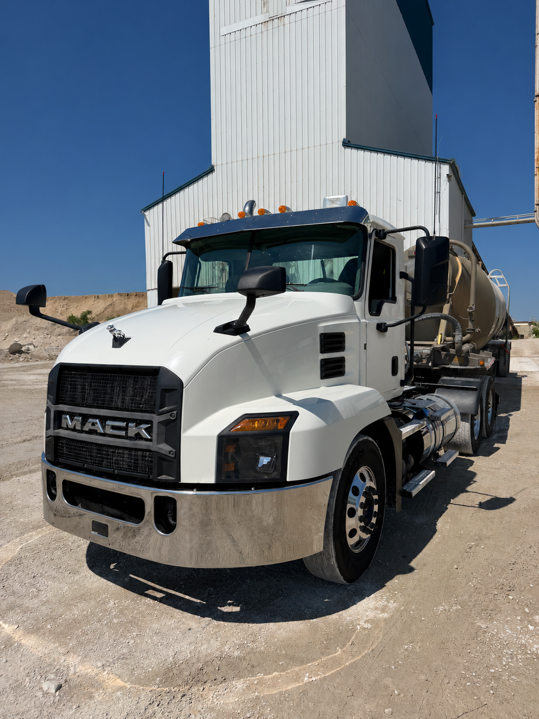 White Mack truck with a bulk trailer beside a construction materials facility
